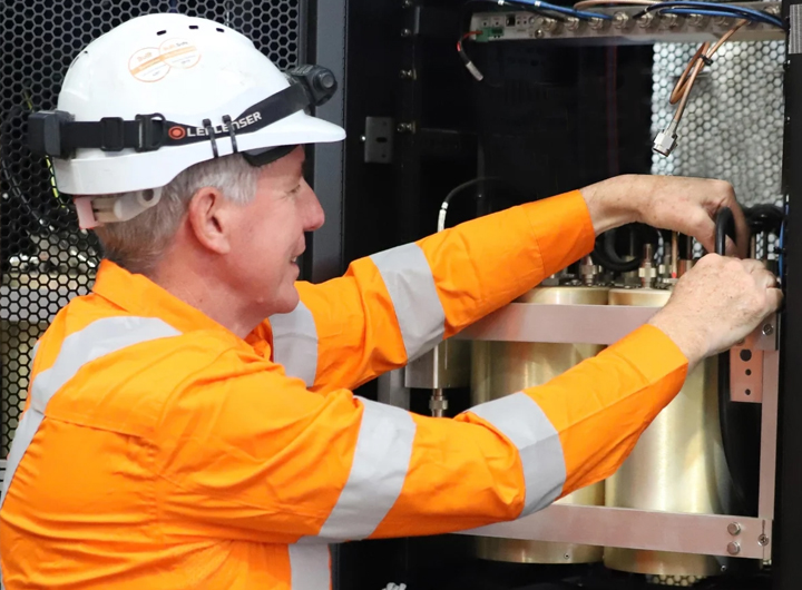 Close up photo of a man in orange hi vis safety long sleeve top and wearing white hard hat is working on an RFI antenna.