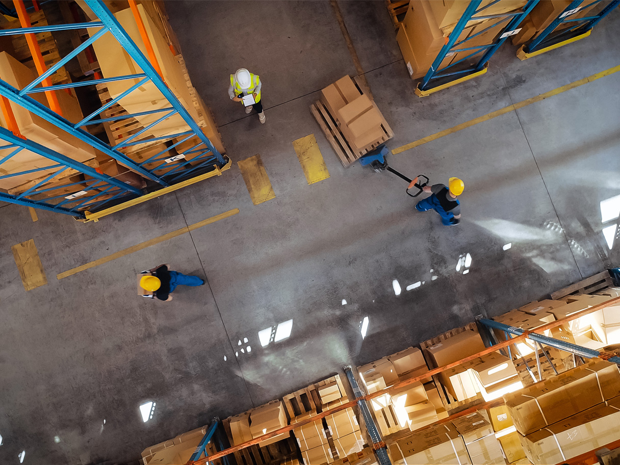 Photo of logistics warehouse with lots of boxes and three people wearing hardhats.