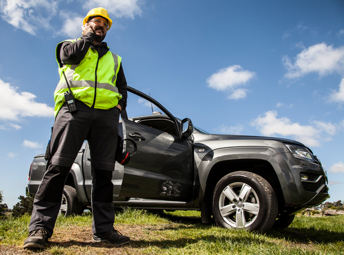 Tait PTToX user on cell phone with LMR radio on hip. He stands in front of a vehicle and he's wearing a yellow hard hat and fluoro work vest.